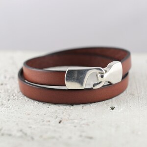 May include: A brown leather bracelet with a silver-colored clasp. The bracelet wraps around twice, showcasing the rich color of the leather. The clasp is a prominent feature, adding a touch of shine to the accessory.