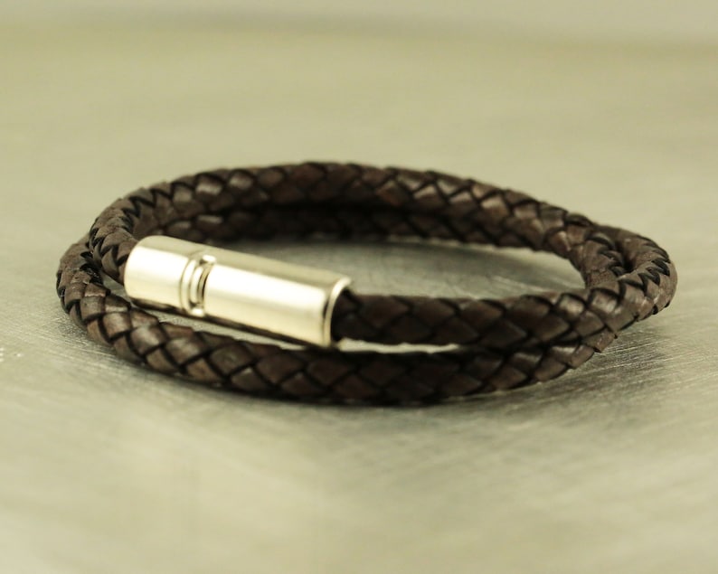 Puede incluir: Un brazalete de cuero marr&oacute;n con un cierre magn&eacute;tico plateado. El brazalete est&aacute; trenzado y tiene un dise&ntilde;o sencillo.