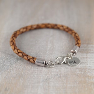 May include: A brown leather braided bracelet with a silver clasp and a silver charm with the letter "K" engraved on it.