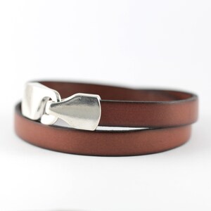May include: A brown leather wrap bracelet with a silver-colored clasp. The bracelet is made of two strands of leather and has a simple, elegant design. The clasp is a rectangular shape.