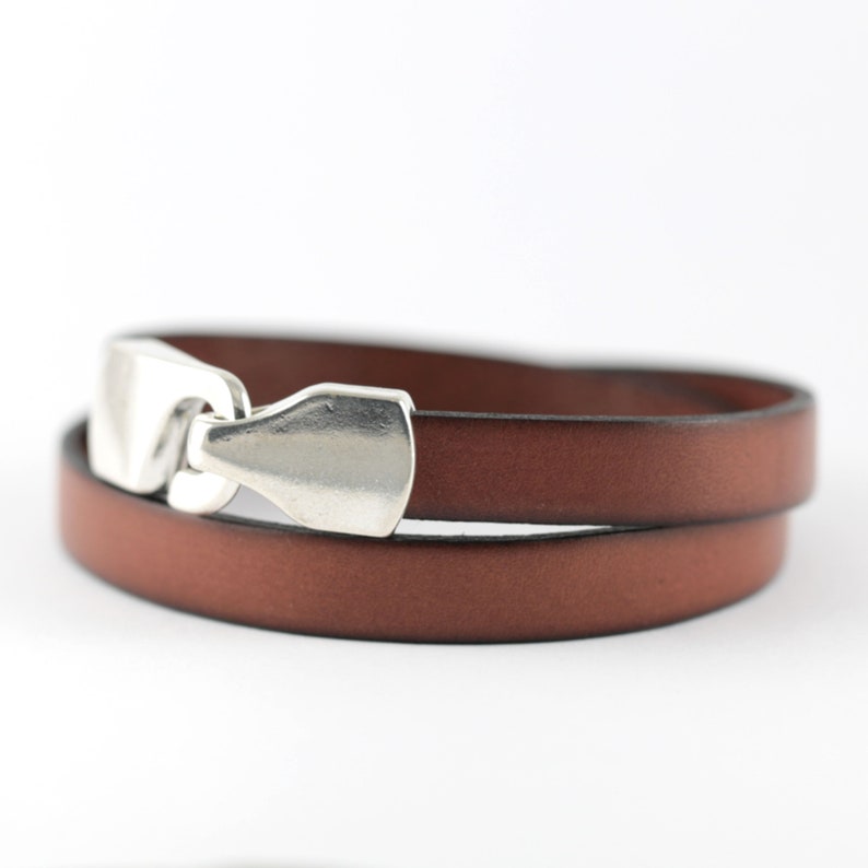 May include: A brown leather wrap bracelet with a silver-colored clasp. The bracelet is made of two strands of leather and has a simple, elegant design. The clasp is a rectangular shape.