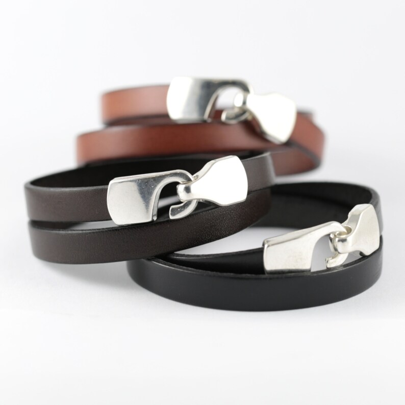 May include: Stack of leather bracelets in brown, dark brown, and black. Each bracelet features a silver-colored clasp. The bracelets are stacked on top of each other, with the top bracelet in brown.