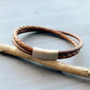 May include: A brown leather bracelet with a silver clasp. The bracelet is braided and has a double layer.