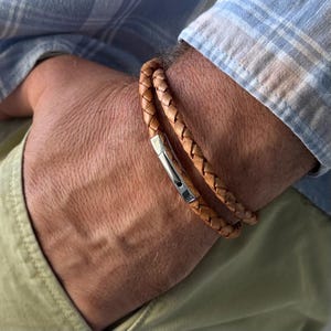Puede incluir: Una pulsera de doble vuelta con tiras de cuero trenzado en color tostado y un cierre rectangular de metal plateado pulido. El cuero presenta una textura natural y el cierre cuenta con un mecanismo de bisagra visible en el lateral de la muñeca.
