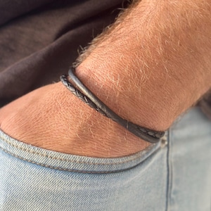 May include: A black leather bracelet with a braided design. The bracelet is worn on a person's wrist.