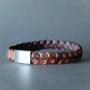 May include: A braided brown leather bracelet featuring a rectangular silver-toned metal magnetic clasp. The leather has a textured, multi-tonal finish ranging from light to dark brown, and the clasp is smooth with a brushed metallic appearance.