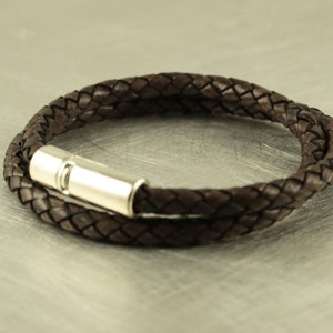 Puede incluir: Un brazalete de cuero marr&oacute;n con un cierre magn&eacute;tico plateado. El brazalete est&aacute; trenzado y tiene un dise&ntilde;o sencillo.