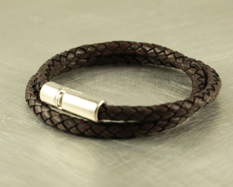 Puede incluir: Un brazalete de cuero marr&oacute;n con un cierre magn&eacute;tico plateado. El brazalete est&aacute; trenzado y tiene un dise&ntilde;o sencillo.