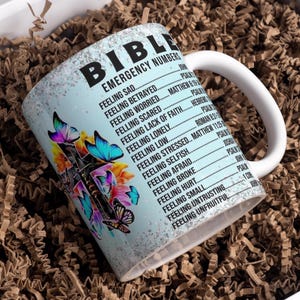 May include: White ceramic mug with a blue background and a cross with butterflies. The mug has the text "Bible Emergency Numbers" and a list of feelings with corresponding Bible verses.