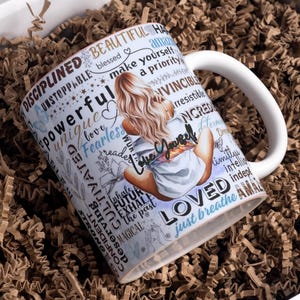 Puede incluir: Taza de cer&aacute;mica blanca con un dise&ntilde;o envolvente que presenta a una mujer sentada con cabello rubio, rodeada de palabras inspiradoras como "poderosa" y "amada". La taza incluye el texto "Love Yourself" y "just breathe".