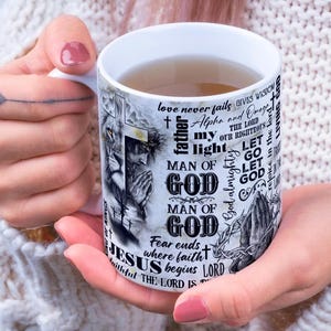 May include: White ceramic mug with a religious theme. The mug features a lion, Jesus, and various religious phrases like "Man of God" and "Jesus". The mug is held by a person.