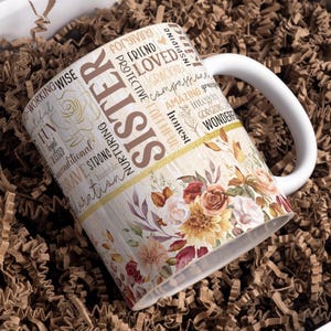 Op de afbeelding: Witte keramische mok met een bloemenpatroon en het woord "Zuster" in bruine letters. De mok is omgeven door bruine verpakkingschips.