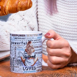 Puede incluir: Taza de cer&aacute;mica blanca con las palabras "Love Yourself" y otras palabras inspiradoras. La taza presenta una ilustraci&oacute;n de una mujer y la sostiene una persona. Un cruas&aacute;n se sostiene en la otra mano.