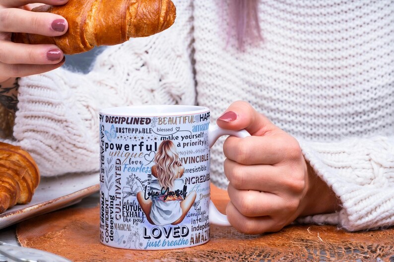 Puede incluir: Taza de cer&aacute;mica blanca con las palabras "Love Yourself" y otras palabras inspiradoras. La taza presenta una ilustraci&oacute;n de una mujer y la sostiene una persona. Un cruas&aacute;n se sostiene en la otra mano.
