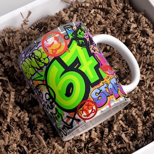 May include: White ceramic mug with a graffiti-style design. The mug features the number "64" in bright green, surrounded by colorful graffiti art, including the word "STAR" and various symbols. The mug has a white handle and is set in a white box with brown packing material.