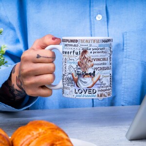 Puede incluir: Una taza de cer&aacute;mica blanca con un dise&ntilde;o envolvente que presenta a una mujer sentada, palabras inspiradoras y la frase "Love Yourself". La taza la sostiene una persona que lleva una camisa azul. Una tableta y pasteles est&aacute;n en el fondo.