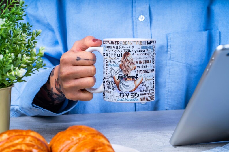 Puede incluir: Una taza de cer&aacute;mica blanca con un dise&ntilde;o envolvente que presenta a una mujer sentada, palabras inspiradoras y la frase "Love Yourself". La taza la sostiene una persona que lleva una camisa azul. Una tableta y pasteles est&aacute;n en el fondo.