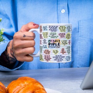 May include: White ceramic mug with a colorful design featuring the text "My Daily Bible Affirmations". The mug is filled with a light brown liquid and is being held by a person's hand.