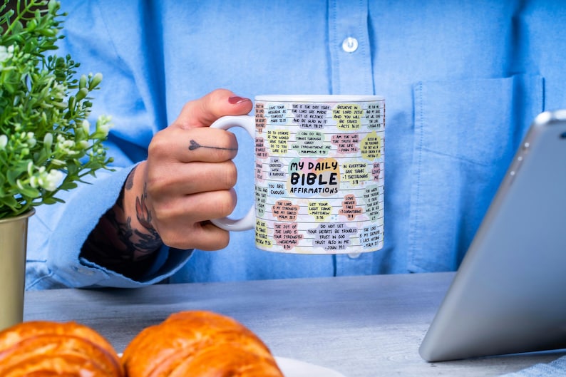 May include: White ceramic mug with a colorful design featuring the text "My Daily Bible Affirmations". The mug is filled with a light brown liquid and is being held by a person's hand.
