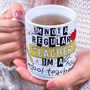 May include: White ceramic mug with the text "I'M NOT A REGULAR TEACHER I'M A COOL TEACHER". The mug features a red apple, a pencil, and a ruler design. The mug is filled with a light brown liquid.