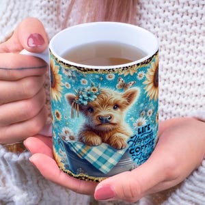 Peut inclure: Mug en c&eacute;ramique blanche orn&eacute; d'une illustration de veau avec une couronne de fleurs, des tournesols et le texte "Life Happens Coffee Helps". Le mug a un fond bleu avec des papillons et des marguerites.