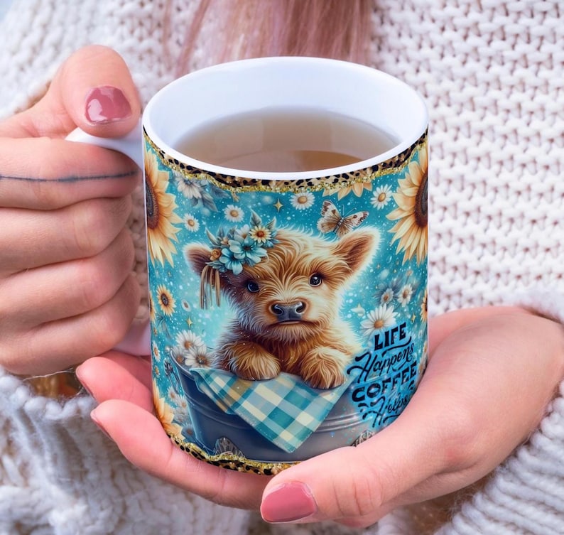 Peut inclure: Mug en c&eacute;ramique blanche orn&eacute; d'une illustration de veau avec une couronne de fleurs, des tournesols et le texte "Life Happens Coffee Helps". Le mug a un fond bleu avec des papillons et des marguerites.