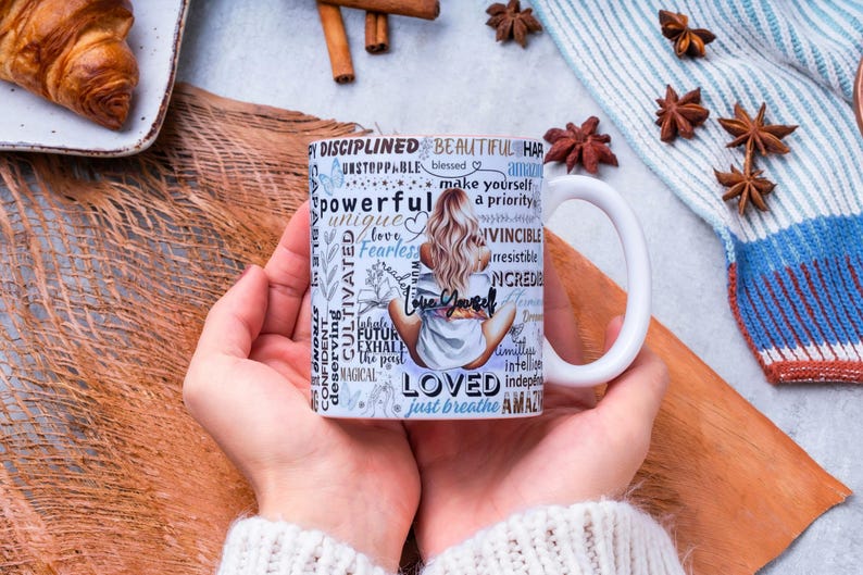 Puede incluir: Taza de cer&aacute;mica blanca con un dise&ntilde;o que presenta a una mujer con cabello rubio y las palabras "Love Yourself" y otras palabras inspiradoras. La taza se sostiene con dos manos, con un fondo de palitos de canela, an&iacute;s estrellado y un cruas&aacute;n.