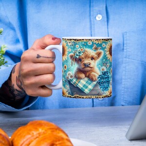 Peut inclure: Mug en c&eacute;ramique blanche avec une illustration de vache des Highlands, des tournesols et le texte &laquo; Life Happens Coffee Helps &raquo;. La tasse a une anse blanche et une bordure dor&eacute;e scintillante. Le fond est bleu clair.