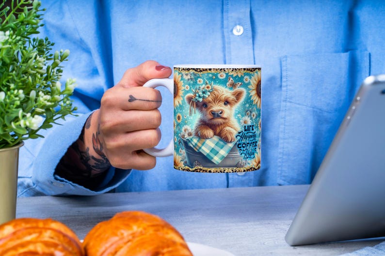 Peut inclure: Mug en c&eacute;ramique blanche avec une illustration de vache des Highlands, des tournesols et le texte &laquo; Life Happens Coffee Helps &raquo;. La tasse a une anse blanche et une bordure dor&eacute;e scintillante. Le fond est bleu clair.