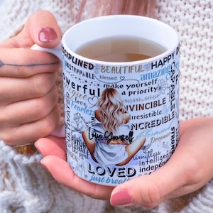 May include: White ceramic mug featuring a woman illustration and motivational words such as "Love Yourself" and "Beautiful". The mug contains a light brown liquid and is held by a person.