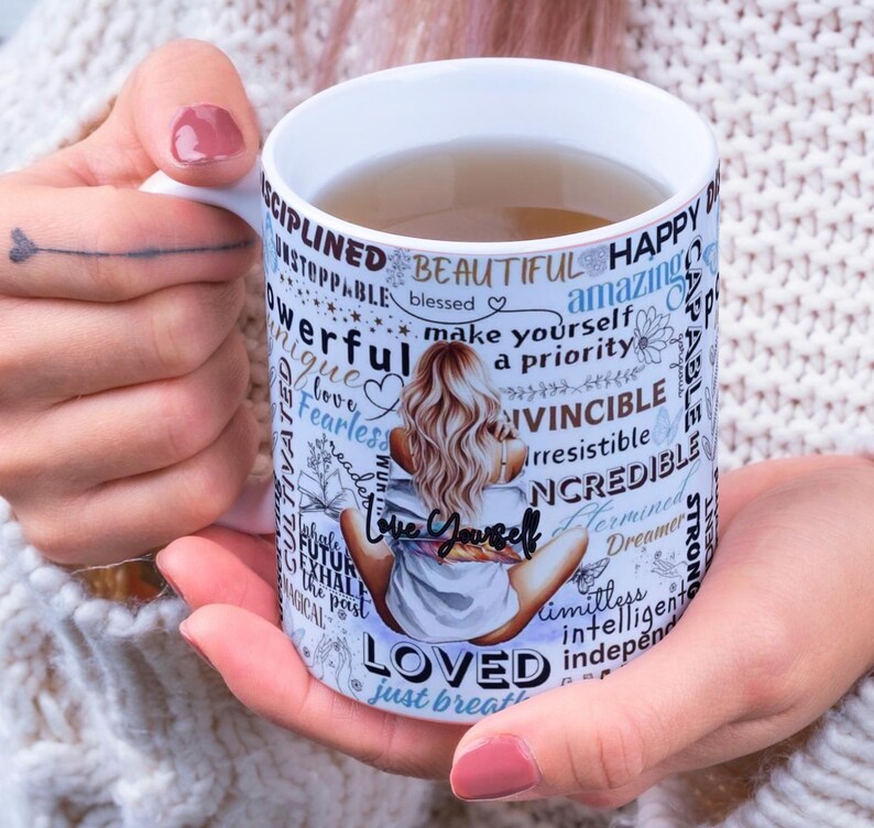 Puede incluir: Taza de cer&aacute;mica blanca con una ilustraci&oacute;n de mujer y palabras inspiradoras como "Love Yourself" y "Beautiful". La taza contiene un l&iacute;quido marr&oacute;n claro y est&aacute; sostenida por una persona.