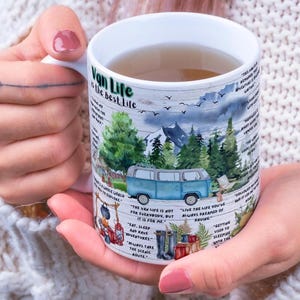May include: White ceramic mug featuring a blue van illustration in a forest setting, with mountains and the text "Van Life is the best Life." The mug is held by a person.