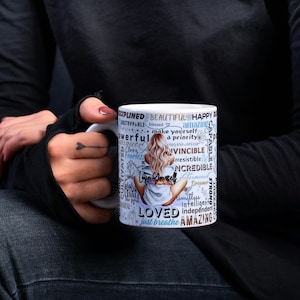 Puede incluir: Taza de cer&aacute;mica blanca con una ilustraci&oacute;n de mujer y las palabras "Amado", "Incre&iacute;ble", "Hermoso" y "Solo respira". La taza es sostenida por una persona que lleva una camisa negra de manga larga y vaqueros oscuros.