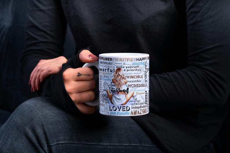 Puede incluir: Taza de cer&aacute;mica blanca con una ilustraci&oacute;n de mujer y las palabras "Amado", "Incre&iacute;ble", "Hermoso" y "Solo respira". La taza es sostenida por una persona que lleva una camisa negra de manga larga y vaqueros oscuros.