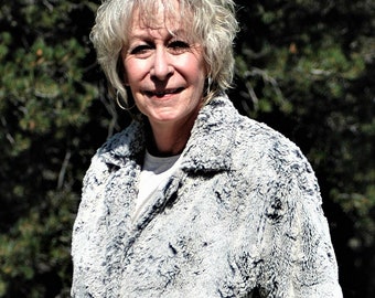 May include: A woman wearing a long, white faux fur coat with a black and white pattern. The coat has a collar and a button closure. The woman is standing in front of a green background.