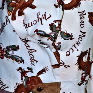 May include: White cotton shirt with a brown and white pattern of cowboys riding horses. The shirt has a brown patch on the bottom right corner and the text "New Mexico" on the right side. The shirt also has the text "Dakota" and "Wyoming" on the front.