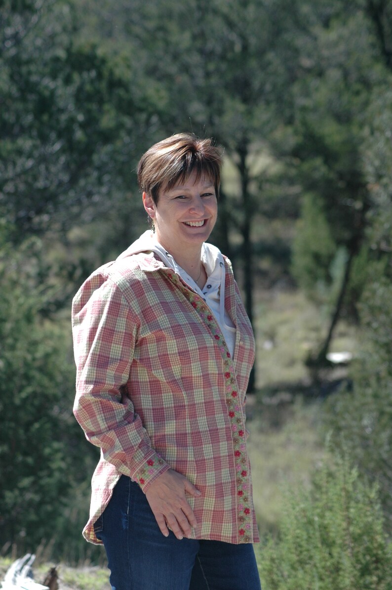 May include: A woman wearing a pink and white plaid shirt with floral embroidery. She is smiling and looking at the camera.