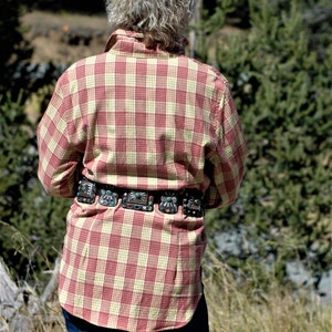 May include: A person wearing a red and yellow plaid shirt with a silver and turquoise belt. The person is standing in a field of tall grass.