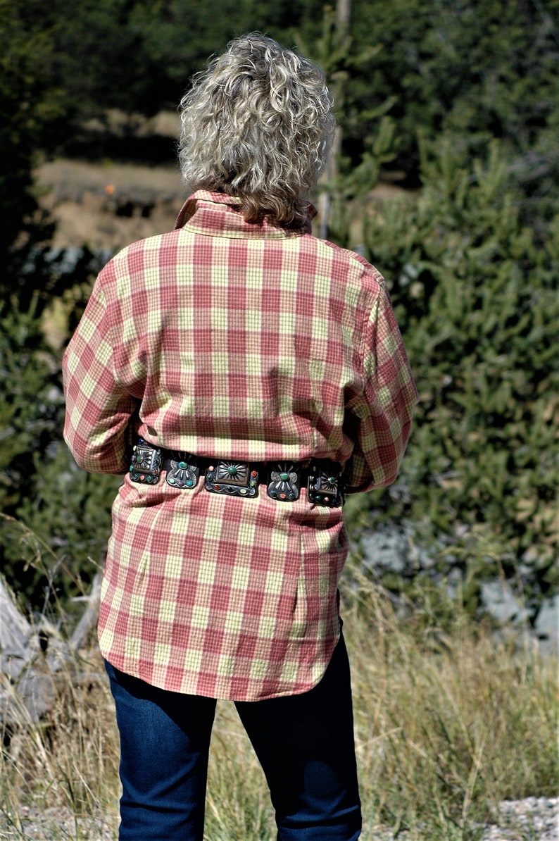 May include: A person wearing a red and yellow plaid shirt with a silver and turquoise belt. The person is standing in a field of tall grass.