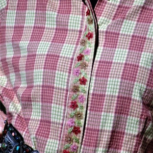 May include: A pink and green plaid shirt with a floral embroidered trim down the front. The shirt is worn with a black leather belt with a silver buckle.