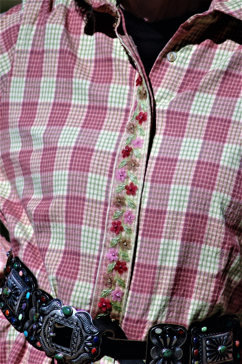 May include: A pink and green plaid shirt with a floral embroidered trim down the front. The shirt is worn with a black leather belt with a silver buckle.