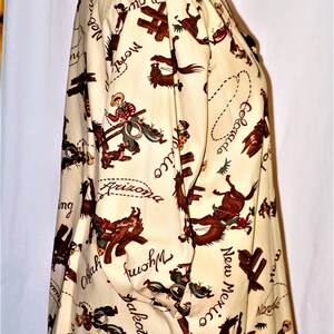 May include: A cream-colored button-up shirt with a brown suede collar. The shirt features a repeating pattern of cowboys riding horses with the names of states written across the fabric. The states include: Montana, Colorado, Nebraska, Wyoming, Dakota, Arizona, Oklahoma, New Mexico, and Texas.