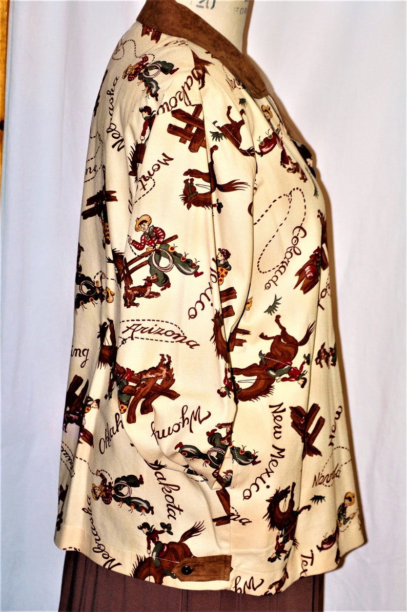 May include: A cream-colored button-up shirt with a brown suede collar. The shirt features a repeating pattern of cowboys riding horses with the names of states written across the fabric. The states include: Montana, Colorado, Nebraska, Wyoming, Dakota, Arizona, Oklahoma, New Mexico, and Texas.