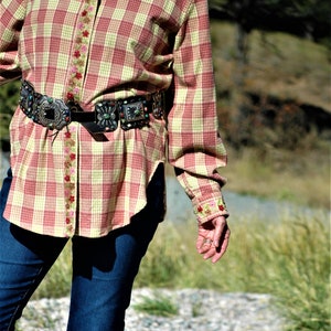 May include: A red and yellow plaid shirt with a floral trim and a silver and turquoise belt buckle. The shirt is worn over blue jeans.