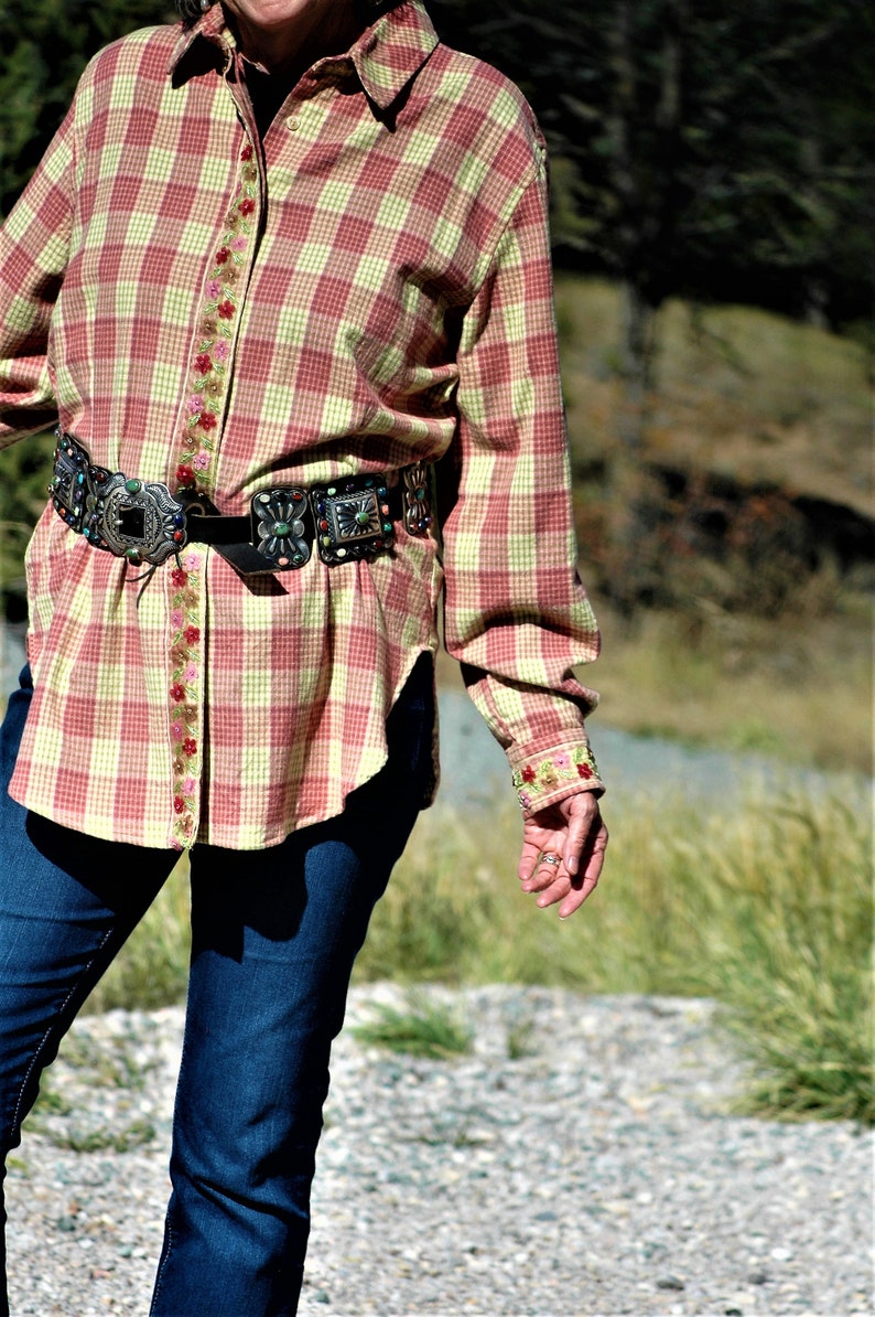 May include: A red and yellow plaid shirt with a floral trim and a silver and turquoise belt buckle. The shirt is worn over blue jeans.