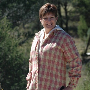May include: A woman wearing a pink and white plaid shirt with floral embroidery. The shirt has a button-down front and long sleeves. She is smiling and looking at the camera.
