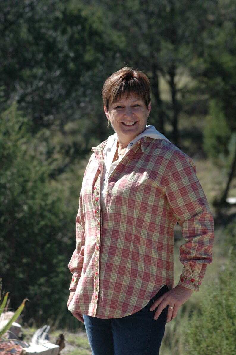 May include: A woman wearing a pink and white plaid shirt with floral embroidery. The shirt has a button-down front and long sleeves. She is smiling and looking at the camera.