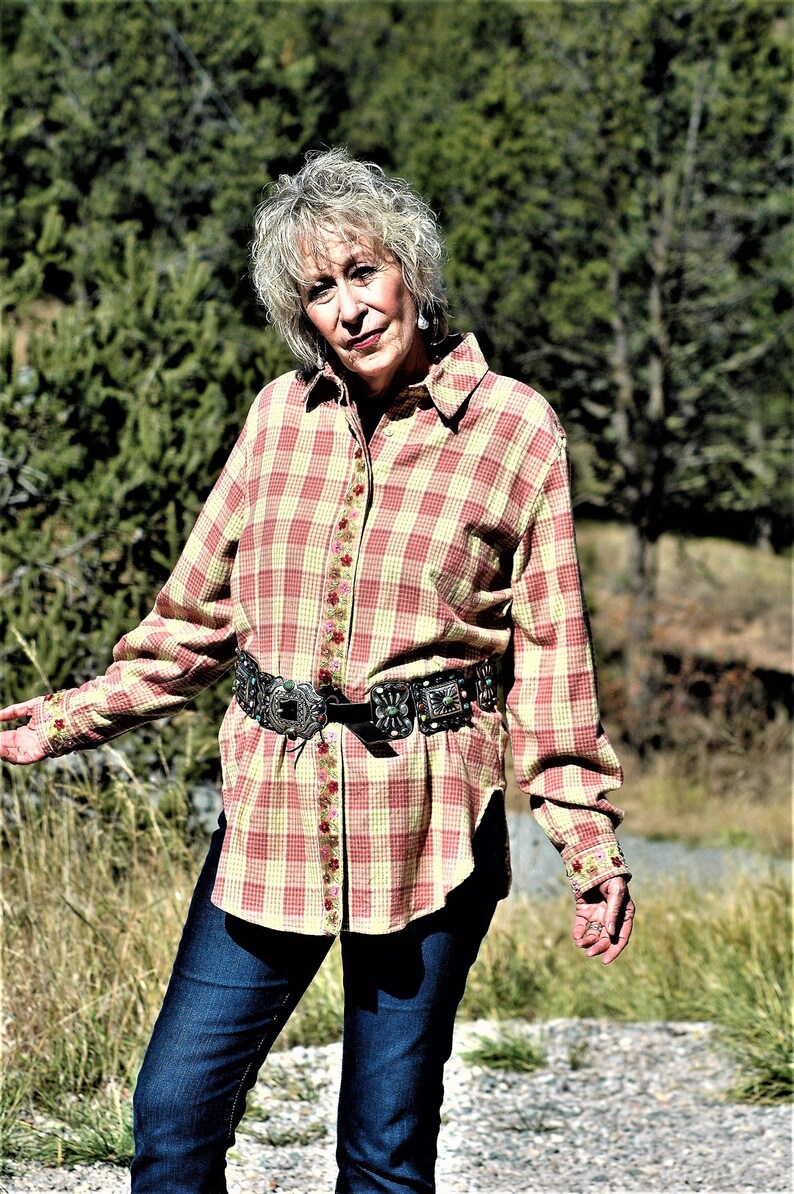 May include: A woman wearing a pink and yellow plaid shirt with a floral trim, a silver belt with turquoise stones, and blue jeans. She is standing in a grassy area with trees in the background.