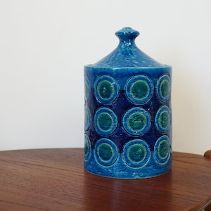 Puede incluir: Un tarro de cerámica azul con tapa, decorado con un patrón repetitivo de círculos verdes. El tarro está sobre una mesa de madera.