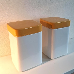 Op de afbeelding: Twee witte keramische containers met vierkante, mosterdkleurige deksels. De containers hebben een rechthoekige vorm met afgeronde hoeken. De deksels zijn glanzend en de containers staan op een wit oppervlak.
